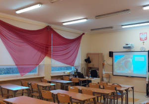Udekorowana sala lekcyjna - rozwieszony na ścianie ogromny czerowny smok oraz stojąca przy tablicy figura Bilba Bagginsa